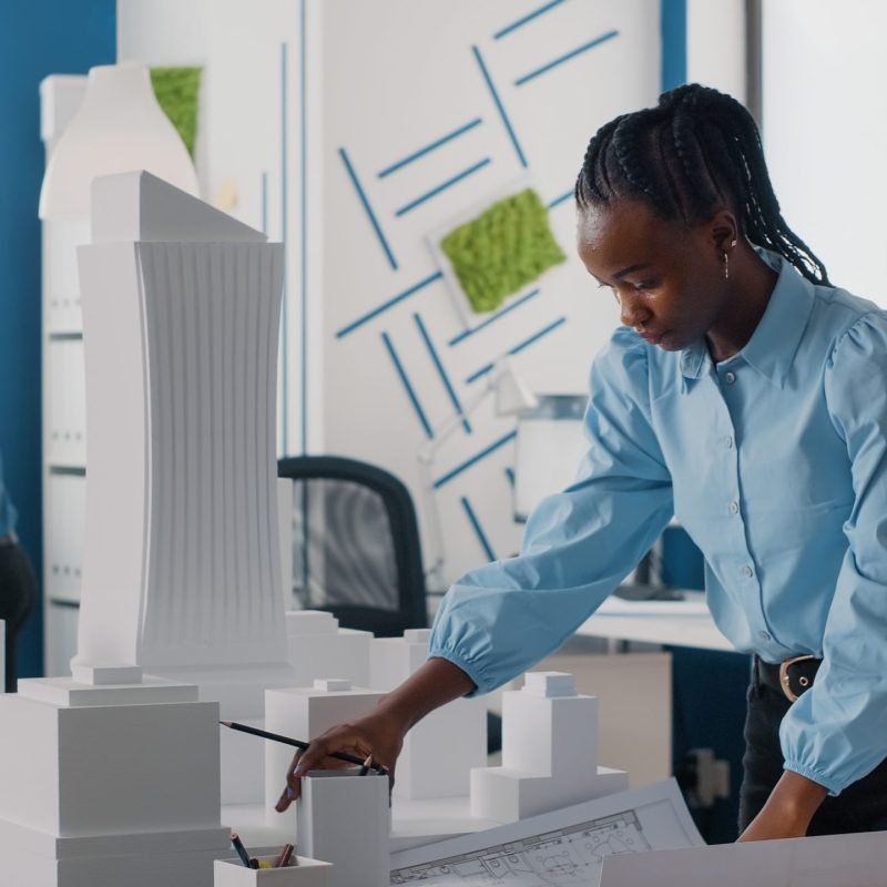 African american woman analyzing blueprints plan and building model to work on architecture. Architect working with maquette and layout print to design urban construction for development.