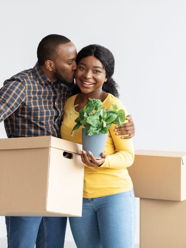 Real Estate Concept. Portrait of happy black couple with cardboard box and plant posing in their new apartment, enjoying moving to own flat, loving husband kissing smiling wife, copy space